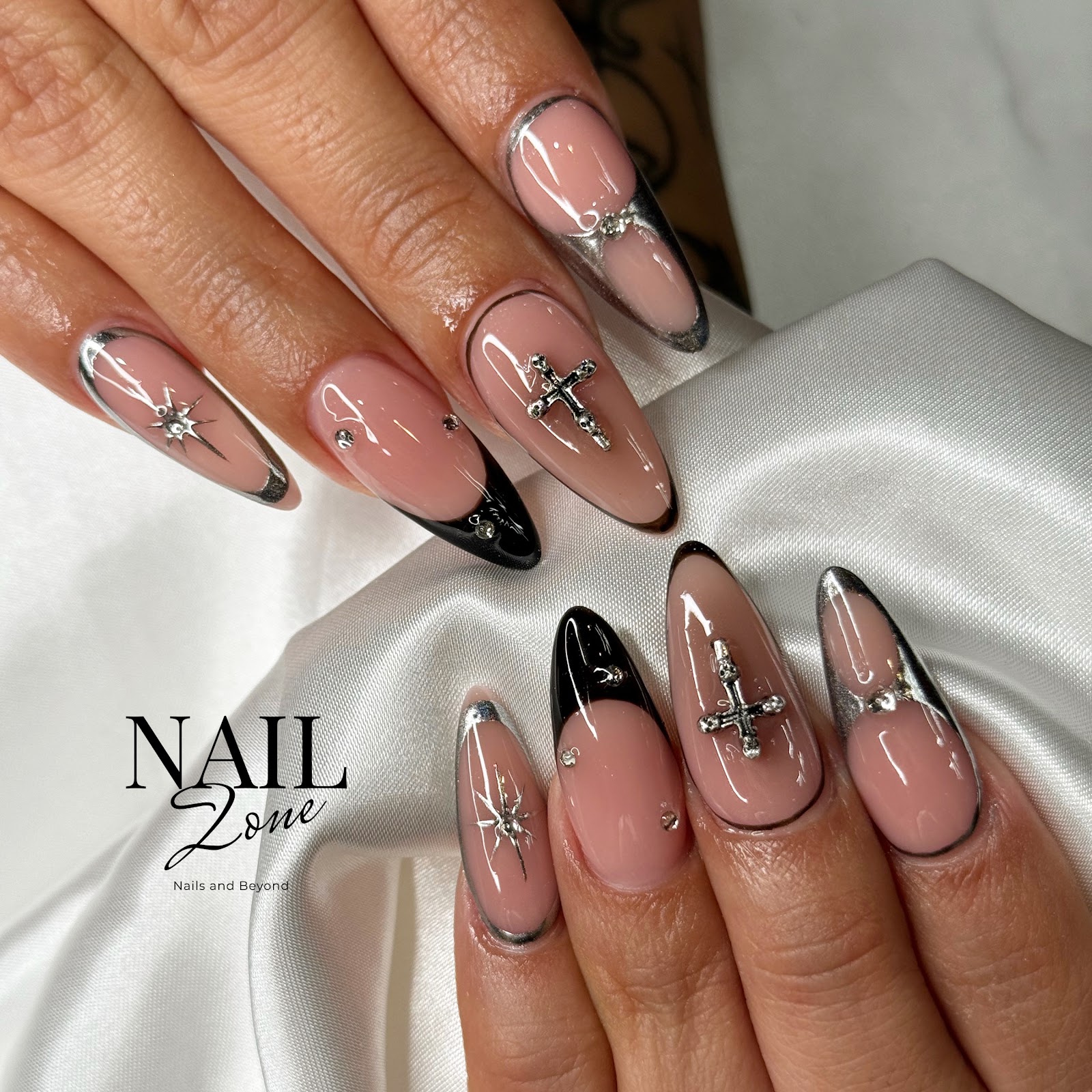 Almond nails in nude, silver-chrome and black tips, finished with cross and star charms.